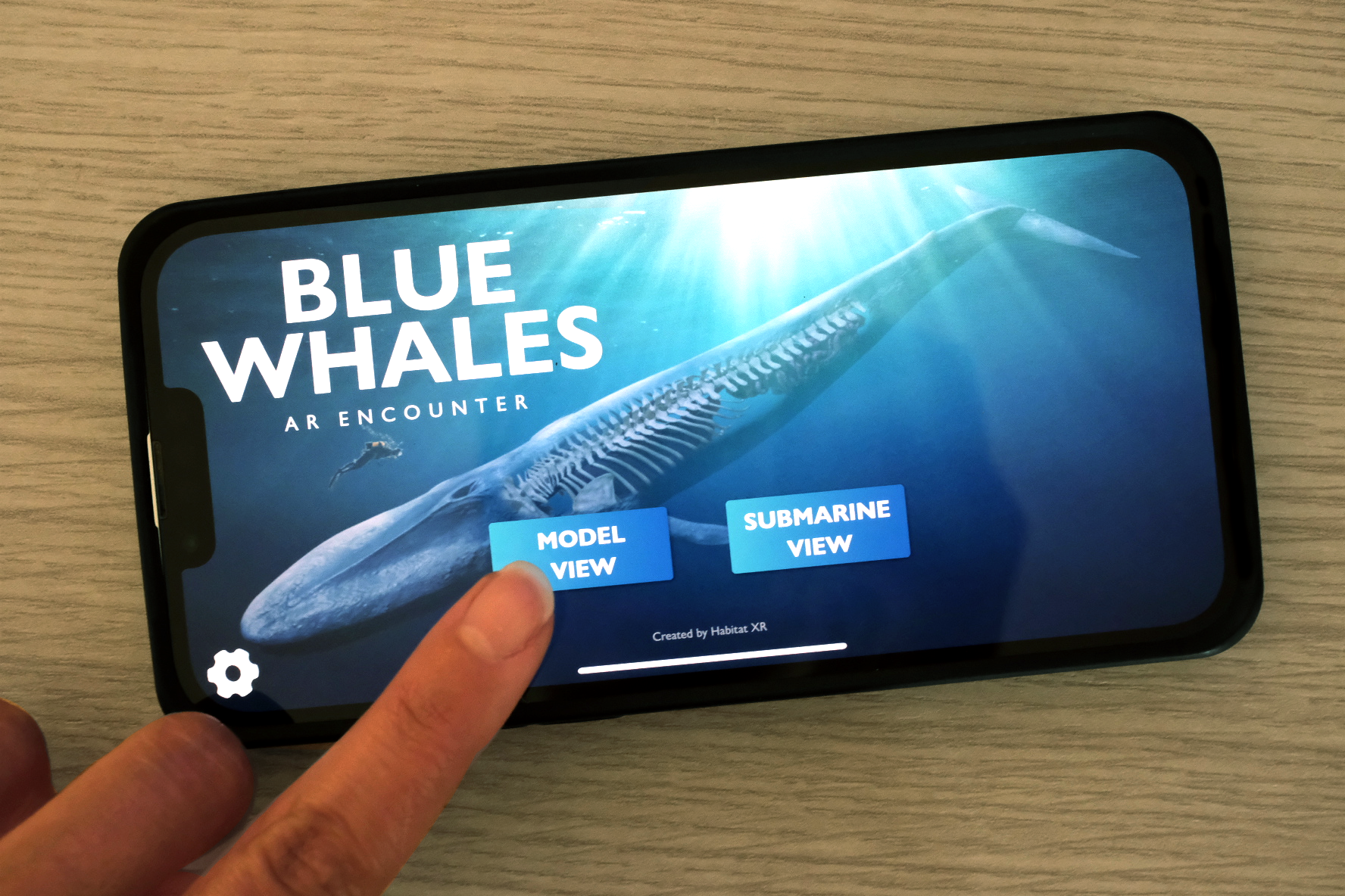 Finger tapping 'Model View' button on a smartphone displaying an AR encounter app featuring a blue whale with visible skeleton underwater.