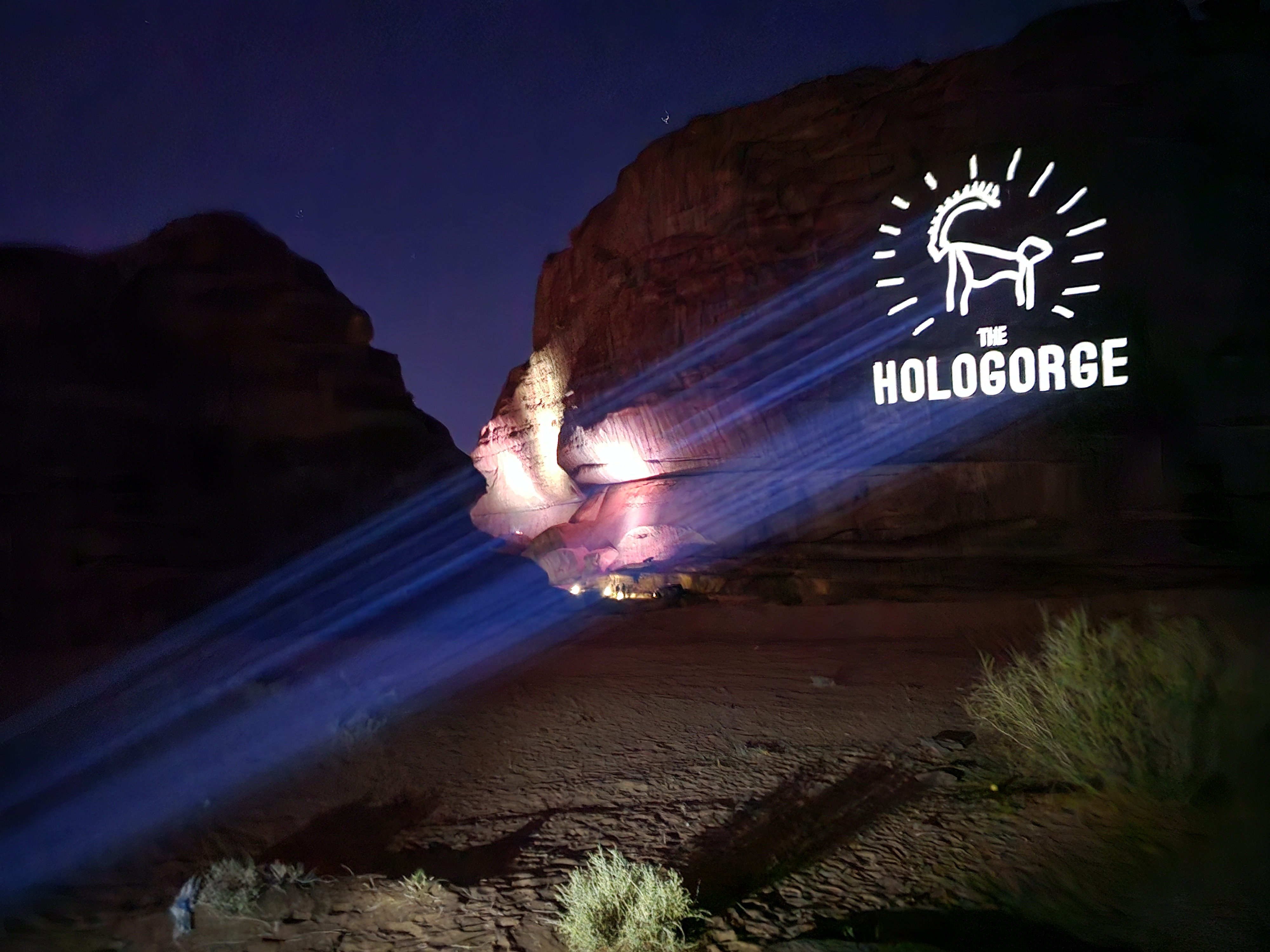 Night scene of desert cliffs illuminated by blue light beams projecting an image of a goat and the words 'The Hologorge'.