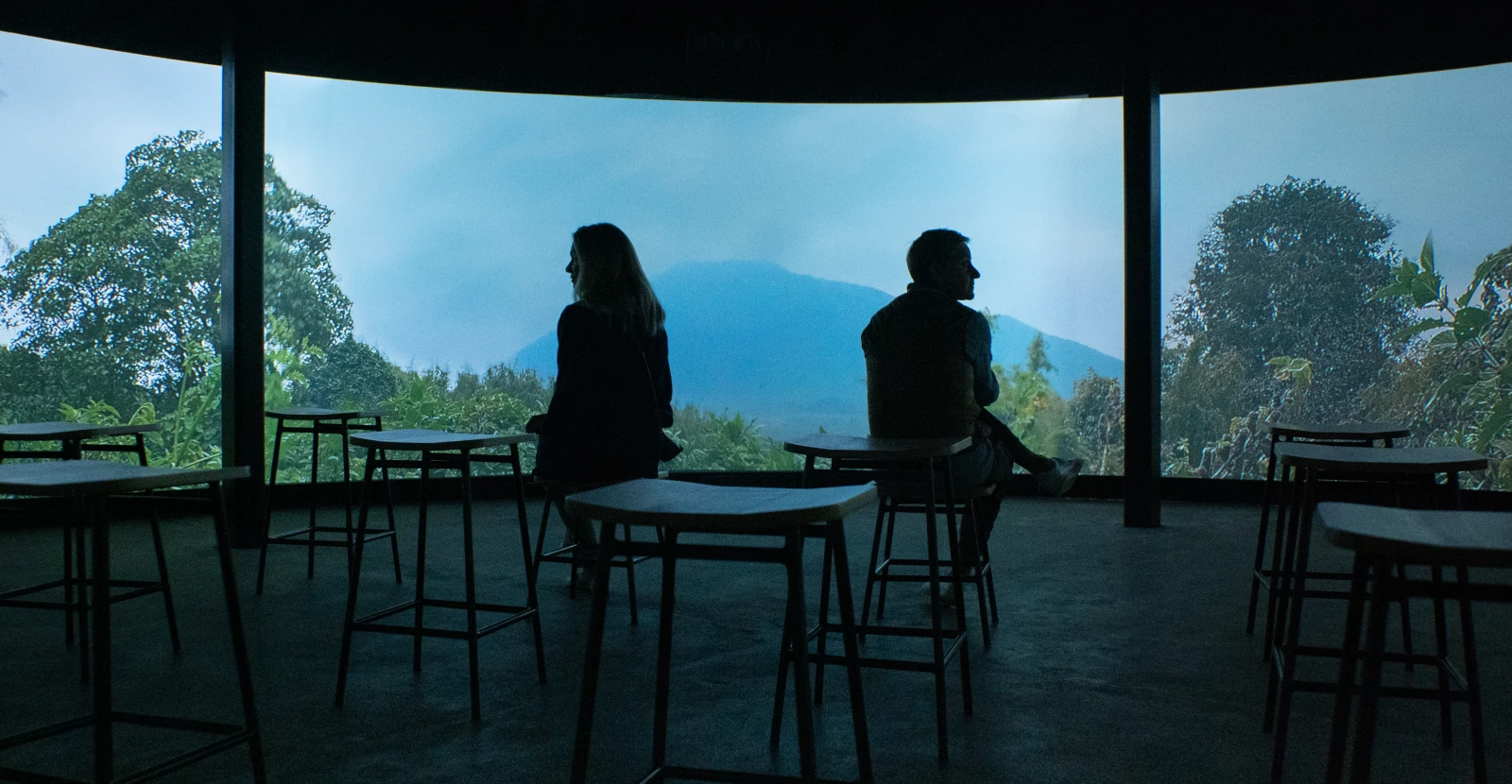 Silhouettes of two people sitting on stools inside a 360 immersive theatre showing a misty mountain in Rwanda